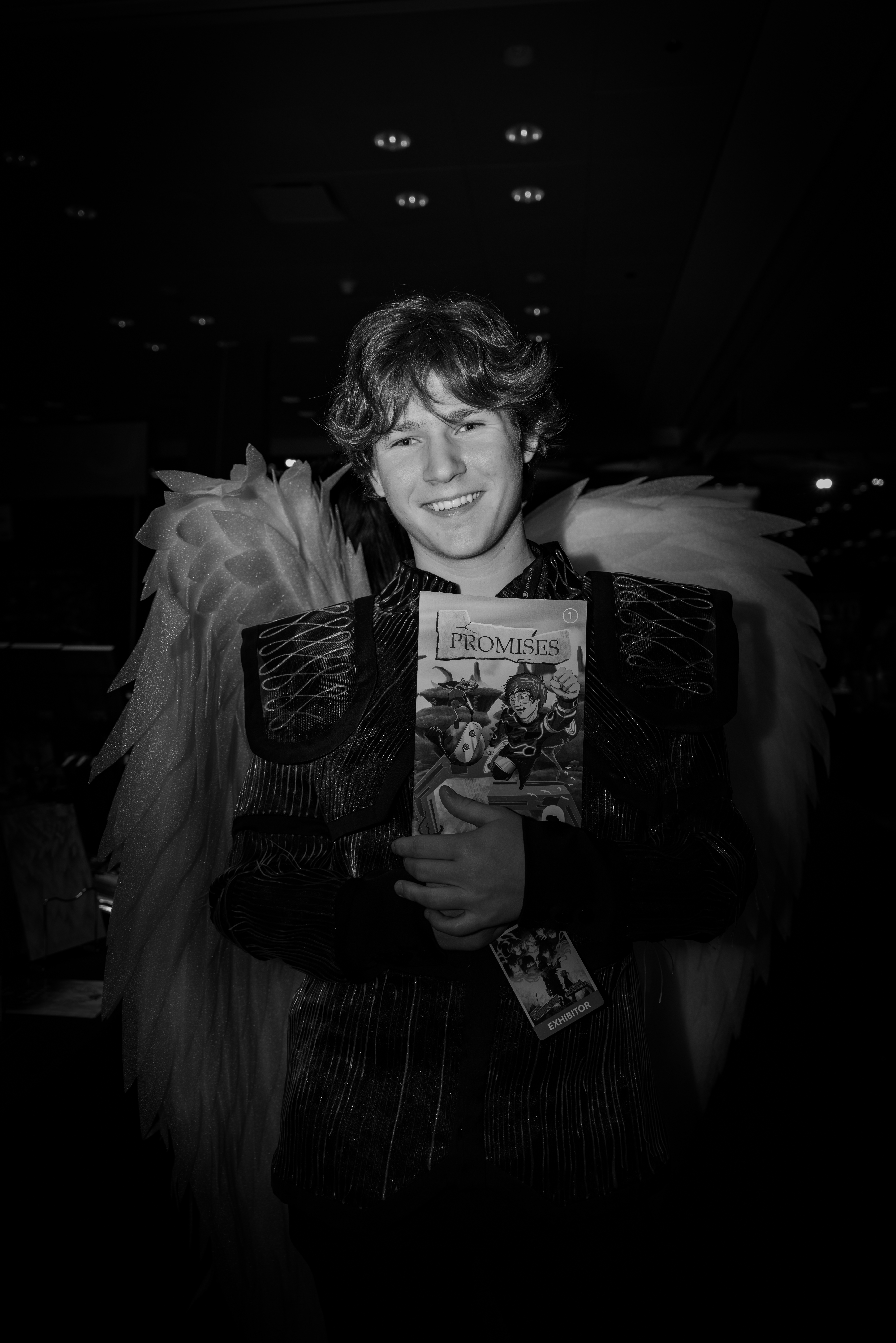 In costume with large feathered wings at New York Comic Con, holding a copy of the comic Promises and wearing an exhibitor badge