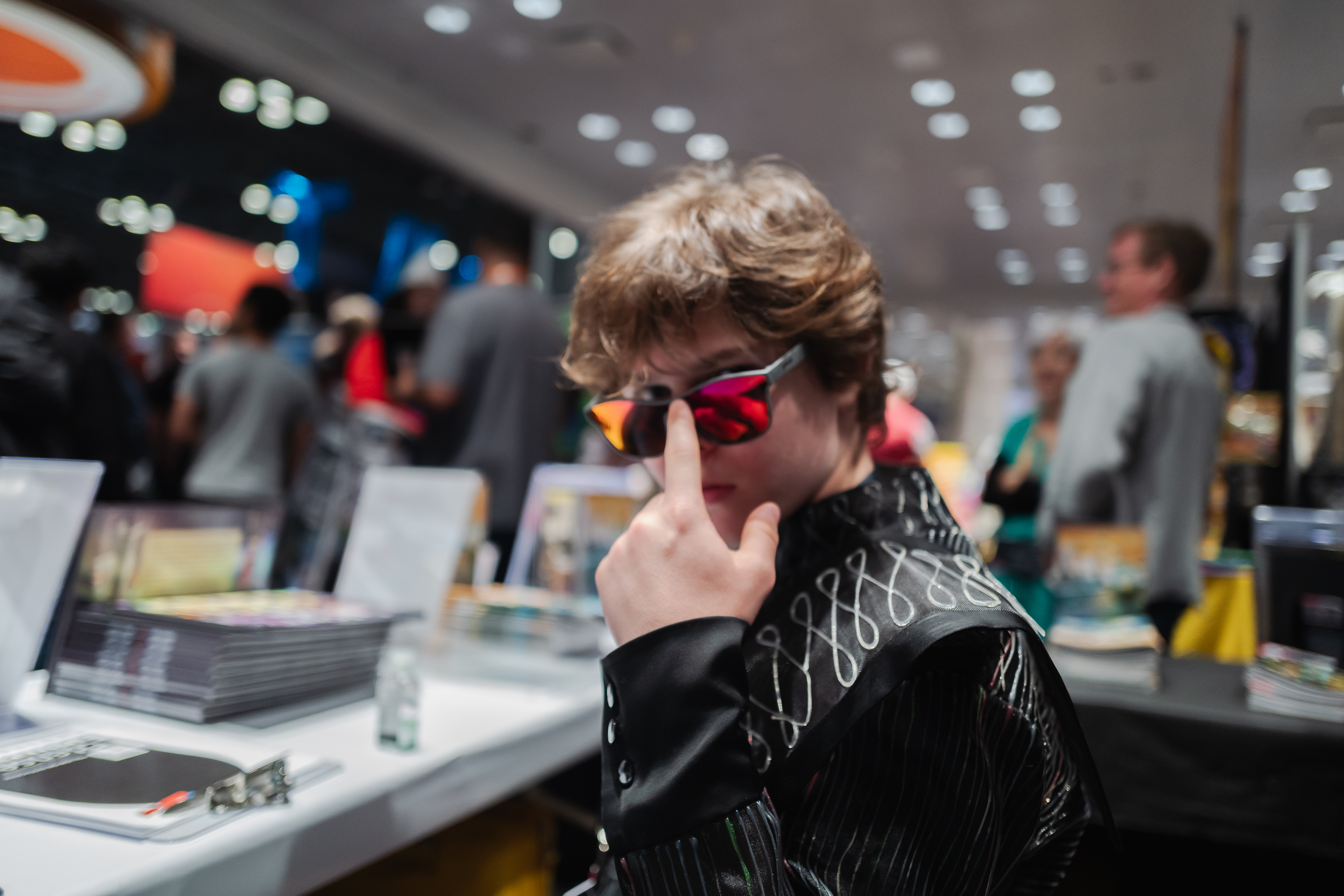 Lowering a pair of red mirrored sunglasses beside a table of books on the Comic Con show floor