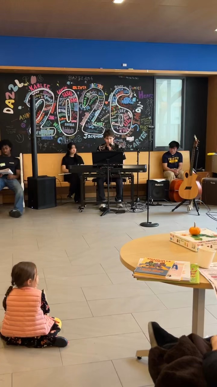 Singing at a keyboard into a microphone while classmates with a guitar and folders sit behind, and a small child watches from the floor