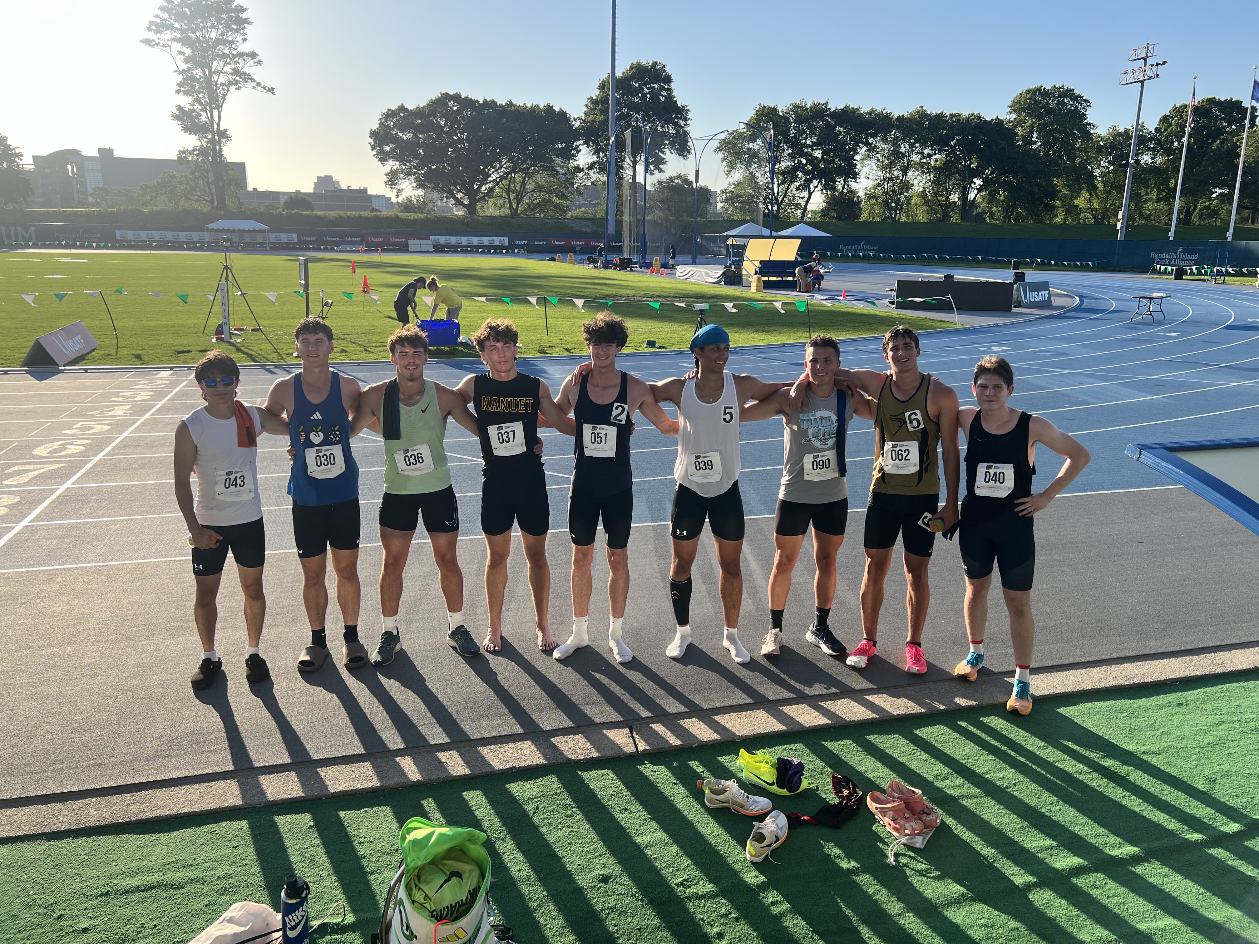 Nine decathletes standing arm in arm across the track at Icahn Stadium in the low evening sun, still wearing their competition bibs
